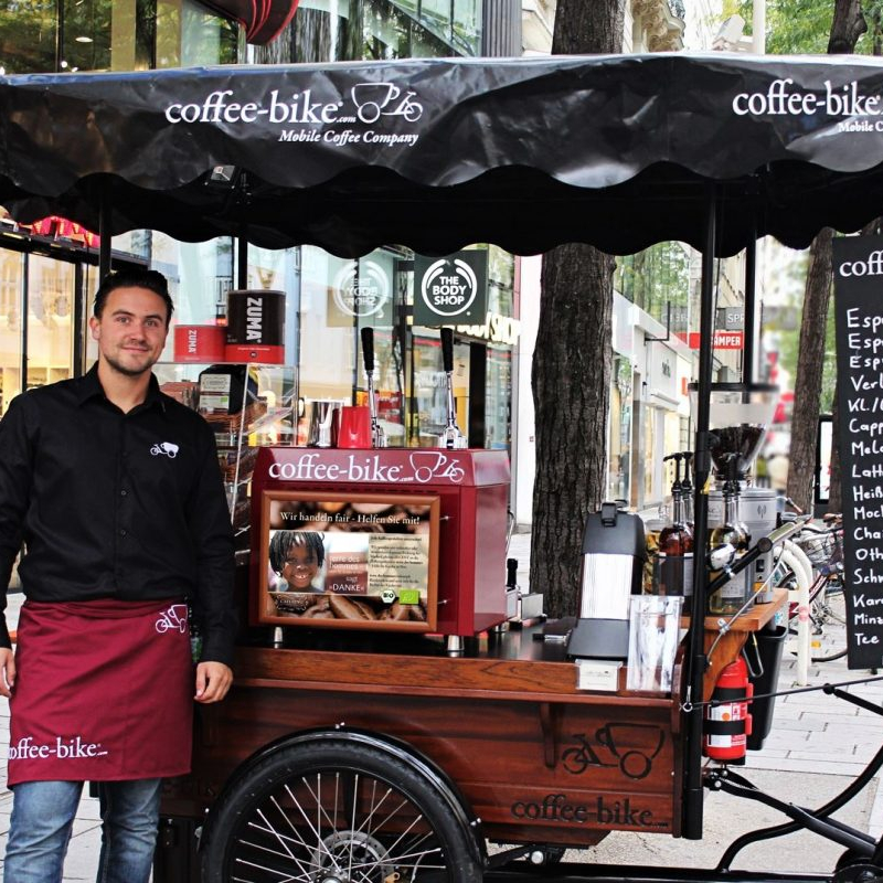Ein Mann im schwarzen Hemd und roter Schürze steht vor dem Coffee-Bike, das sich in einer Innenstadt befindet.
