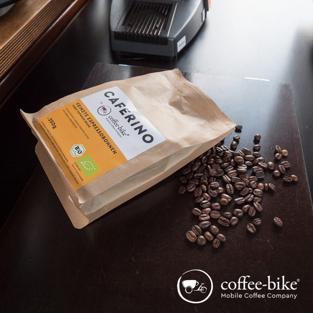 Eine offene Packung Caferino Espressobohnen liegt auf schwarzem Hintergrund, davor vereinzelt Bohnen, unten rechts das weiße Coffee-Bike Logo
