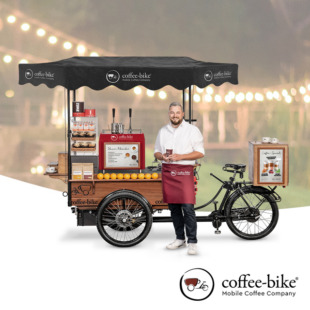 A barista stands in front of a Coffee-Bike, in the background lights, Coffee-Bike Logo