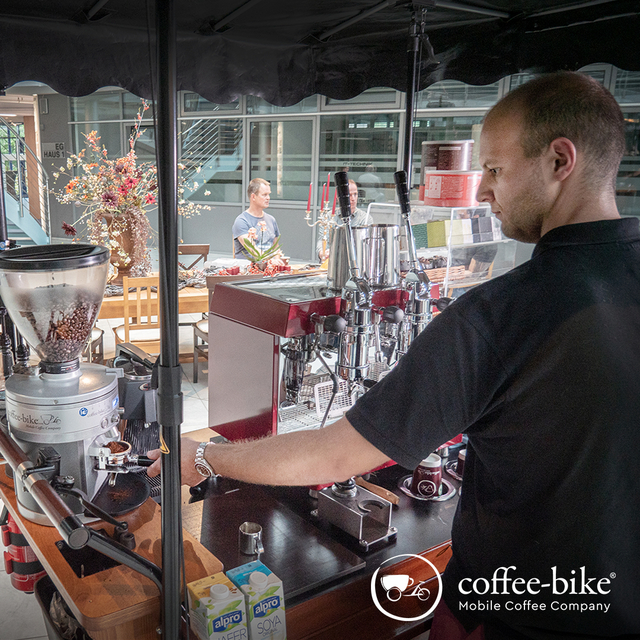 Ein Mann im schwarzen T-Shirt steht am Coffee-Bike und mahlt Bohnen