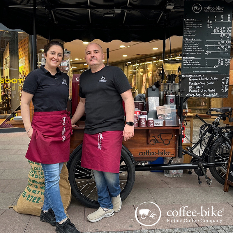 Two people in Coffee-Bike clothing stand in front of the Coffee-Bike and smile.