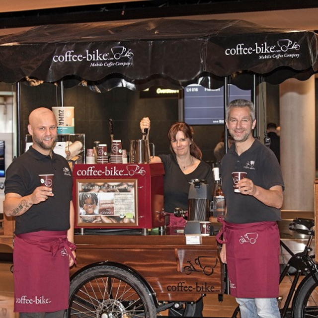 Two men stand in front of the Coffee-Bike with a cup in their hands and a woman stands at the lever of the portafilter machine behind the Coffee-Bike