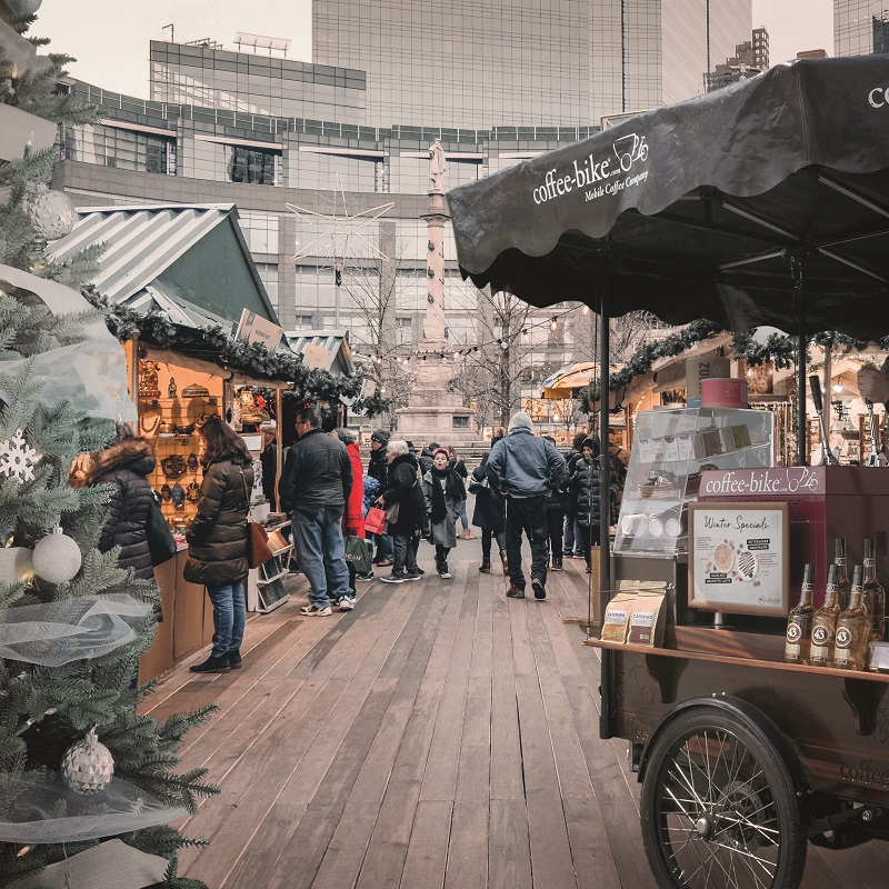 A Christmas market with stalls and people, a cut fir tree on the left side and a cut Coffee-Bike on the right side