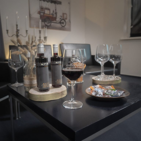 On a black table in front of a candlestick are two bottles of red wine, in front of it a filled red wine glass next to it a candy bowl and behind it two empty wine glasses