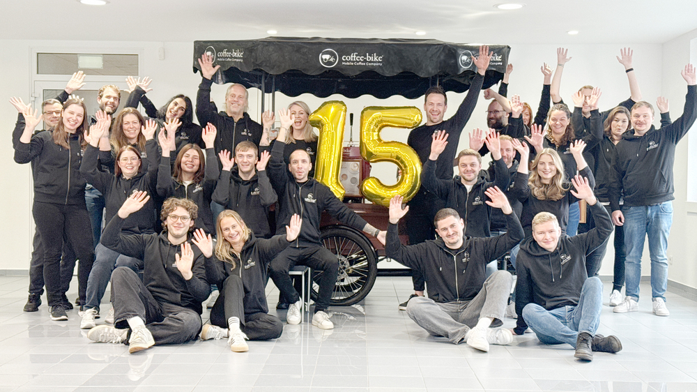 Coffee-Bike Mitarbeiter jubeln vor dem Coffee-Bike und halten eine goldene 15 als Luftballons