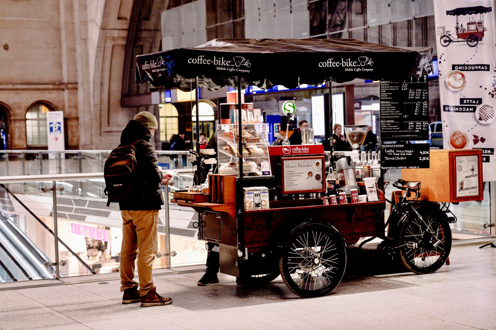 Ein Mann steht links von einem vollausgestatteten Coffee-Bike, rechts ein Flagbanner