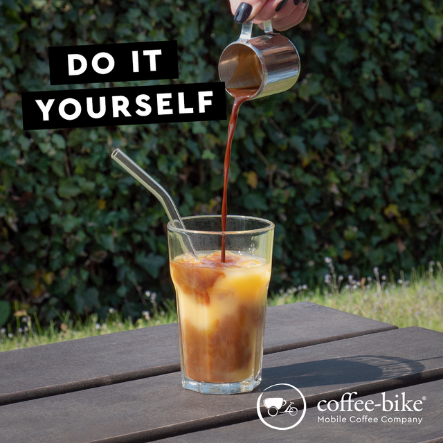 A hand pours espresso with a stainless steel pot from above into a glass with straw and orange juice, above it in black bars is written in white letters Do it yourself, right below white logo