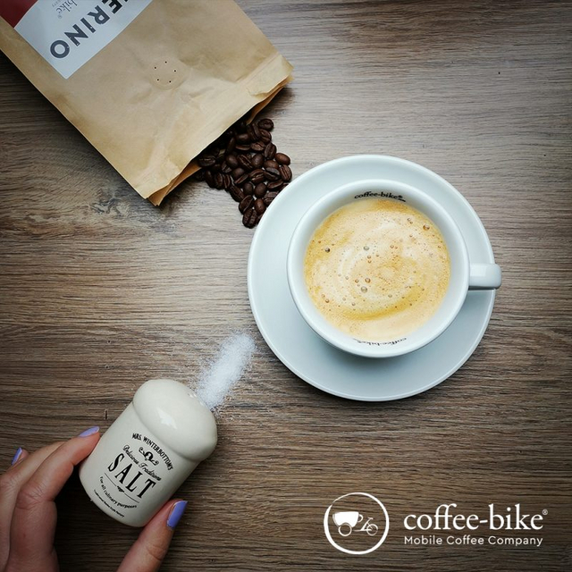 On a brown background is an opened bean packet, a hand is sprinkling some salt with a salt shaker, in between is a white cup with coffee, on the bottom right is the Coffee-Bike logo