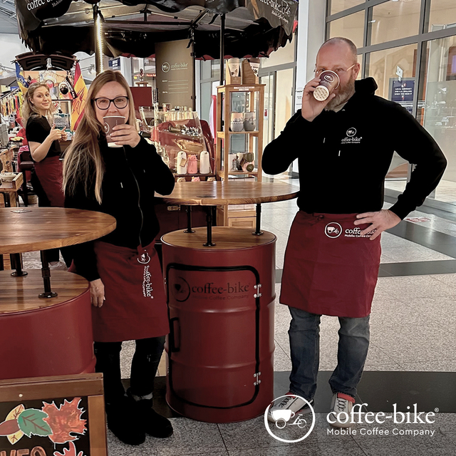 Eine Mann und eine Frau in Coffee-Bike Kleidung und roter Schürze trinken aus Coffee-Bike Bechern, während sie neben Stehtischen stehen, dahinter das Coffee-Bike und eine Barista, rechts unten weißes Coffee-Bike Logo