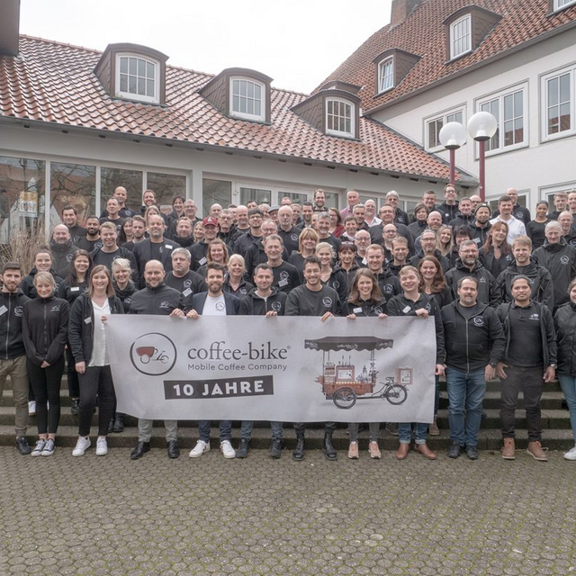 Coffee-Bike Mitarbeiter und Franchisepartner stehen mit einem Jubiläumsbanner für 10 Jahre Coffee-Bike vor dem Haus der Jugend in Osnabrück