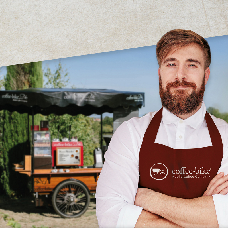 A man with beard, white shirt, red apron and folded arms is placed in front of blurred Coffee-Bike in the background, above is gray stone background