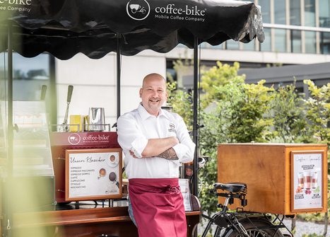 Partenerul de franciză cu tricou și șorț Coffee-Bike zâmbește și se sprijină de partea din față a barului mobil de cafea 