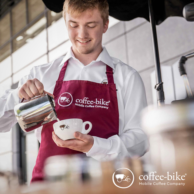 Ein Mann mit weißem Hemd und roter Schürze steht am Coffee-Bike und gießt Milch in eine Tasse ein.