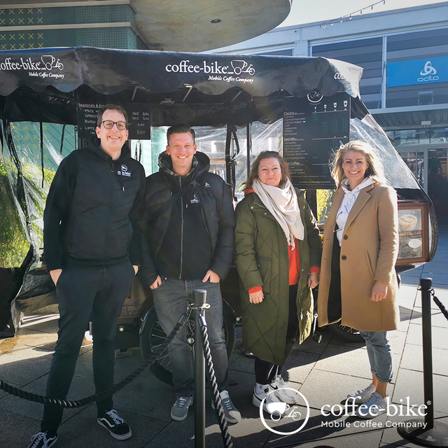 On the left are two men and on the right two women standing in front of the Coffee-Bike, on the bottom right white Coffee-Bike logo