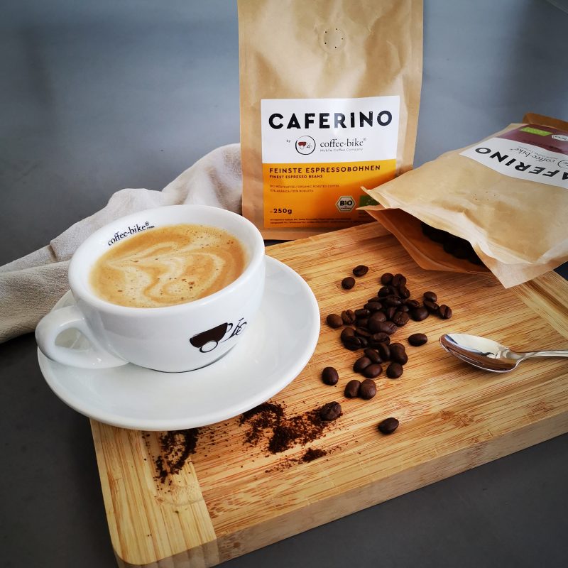 Eine Coffee-Bike Tasse mit Kaffee, eine stehende Espressobohnenpackung, eine geöffnete liegende Kaffeebohnenpackung, vereinzelte Bohnen und ein Löffel sind auf einem Holzbrett verteilt.