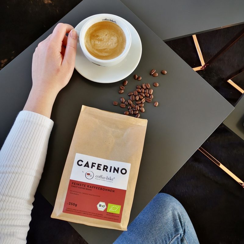 A left arm lies on a table and touches a white coffee cup. In front of it lies a Caferino coffee bean package and single beans.