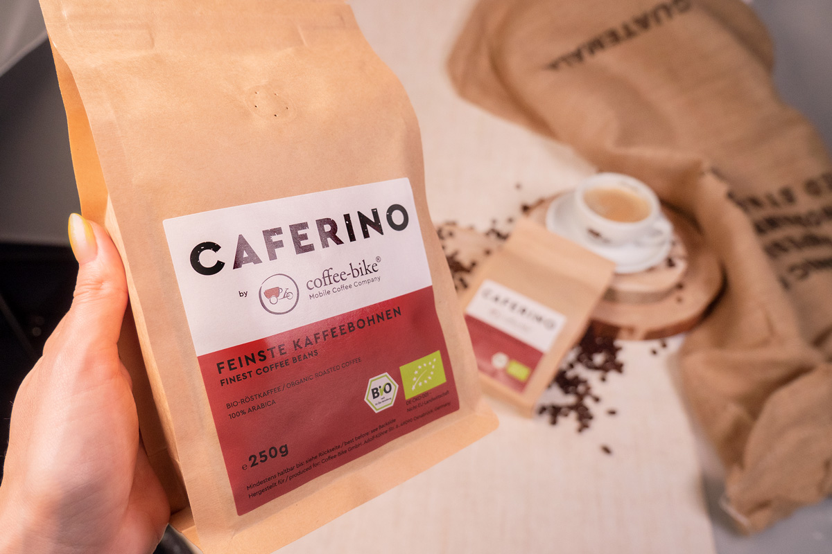 Hand holding coffee package in the foreground in front of a lying Caferino package and a white cup of coffee