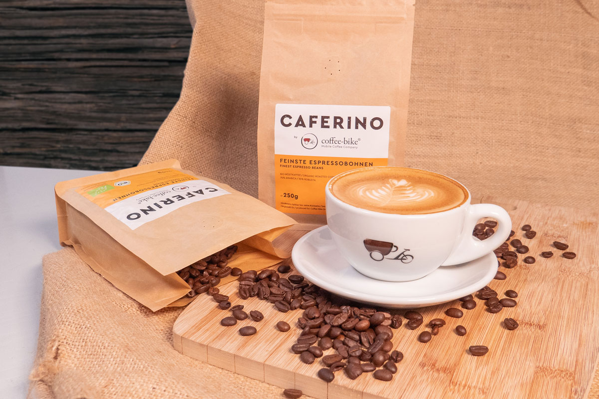 Gefüllte Coffee-Bike Cappuccino Tasse vor zwei Caferino Bio-Espressobohnenpackungen, aus denen Bohnen fließen