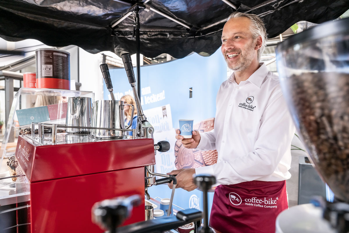 Coffee-Bike Barista hält Kaffeebecher mit myChoco Logo hinter der mobilen Kaffeebar in der Hand und lächelt in Kundenrichtung