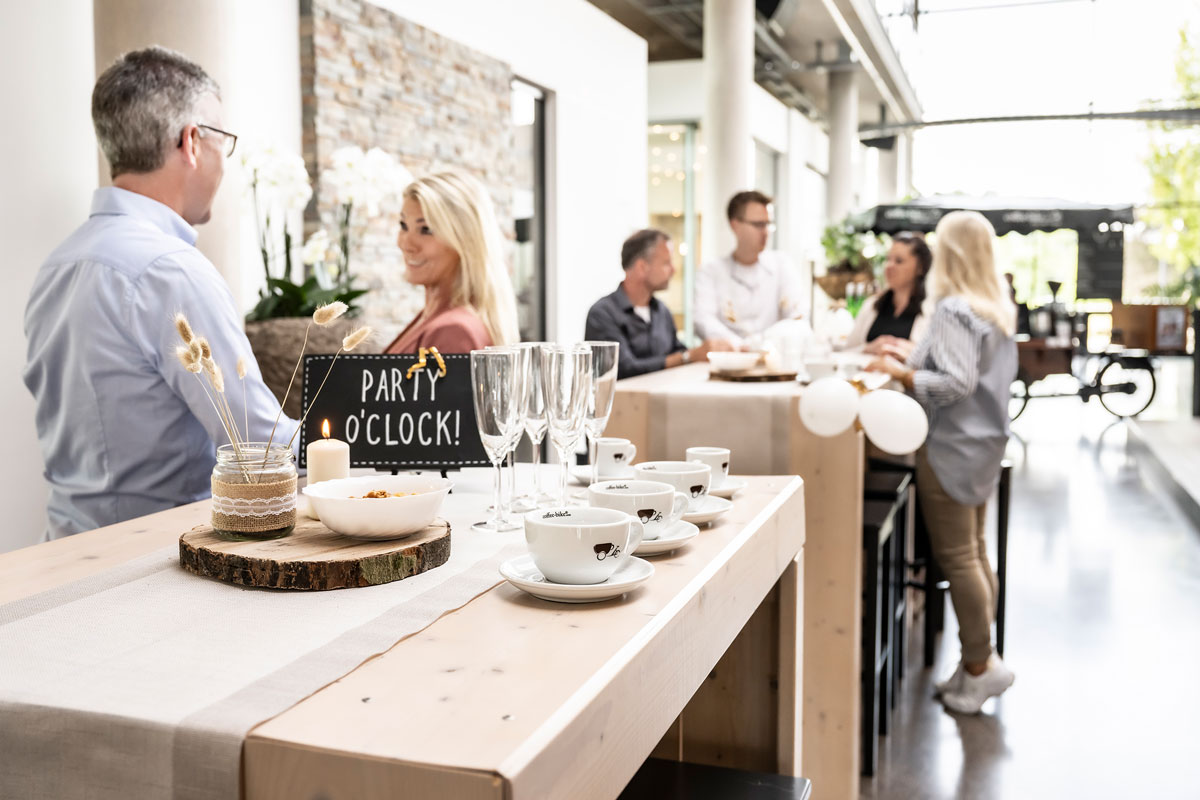 Zona de recepție a unei clădiri corporative cu șase persoane la două mese de bar aranjate în mod festiv, purtând conversații în fața unui bar de cafea mobil