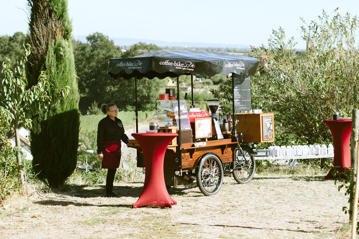 Coffee-Bikerin steht hinter ihrer mobilen Kaffeebar zwischen Wiesen, die dekorativ geschmückt und mit Stehtischen ausgestattet sind
