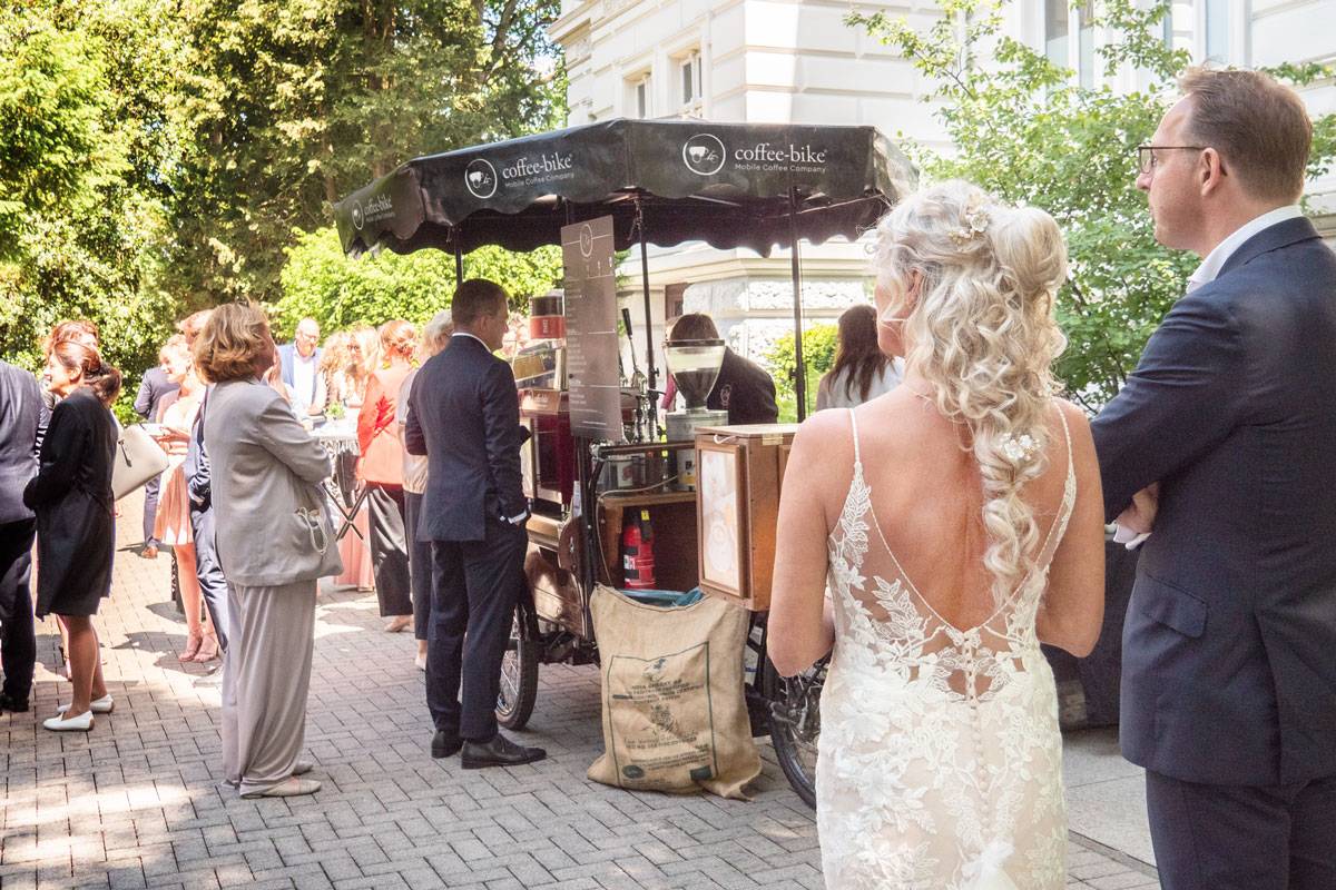 In the greenery a bride and groom with guests stand in front of the Coffee-Bike with two baristas and look in at the drinks table