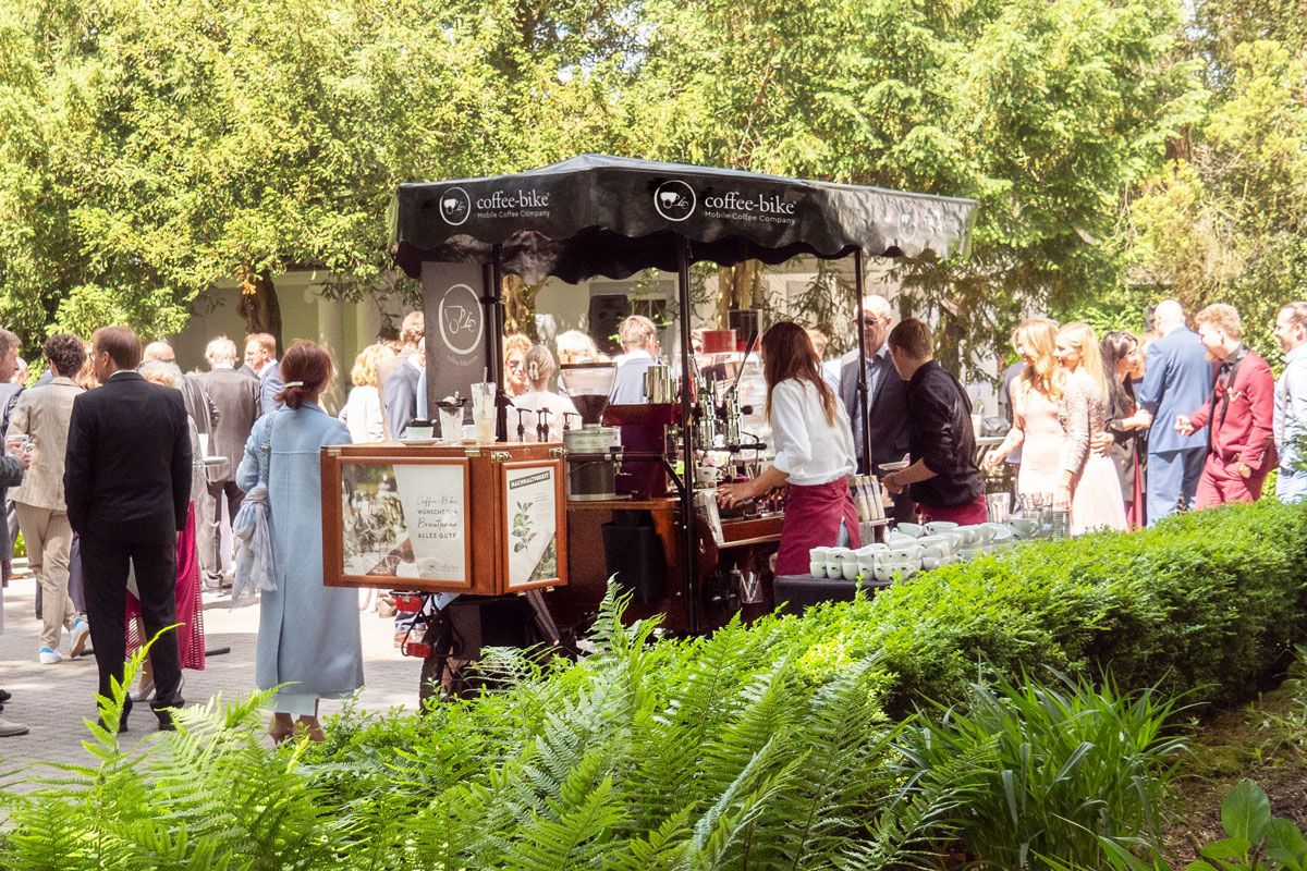 Mobile coffee bar in green outdoor area between festively dressed wedding party on barista perspective