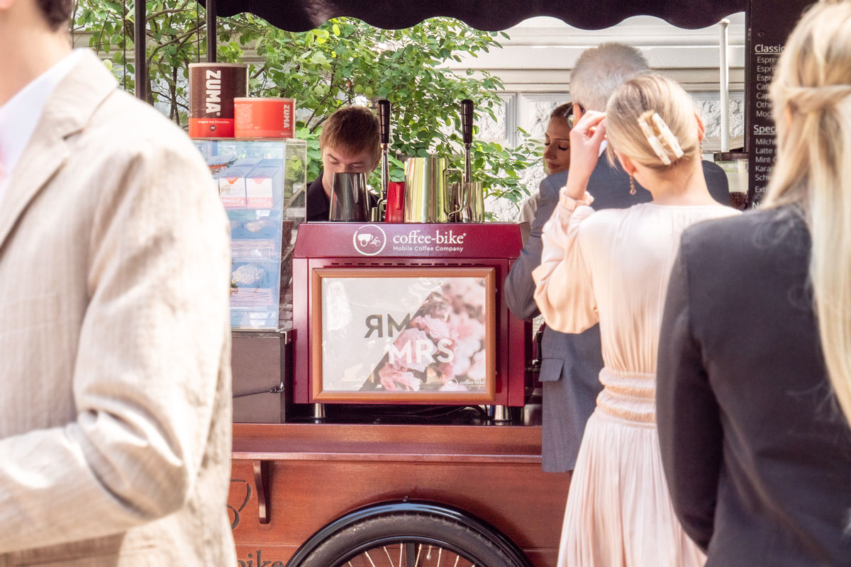 Hochzeitsposter an der Siebträgermaschine des Kaffeefahrrads im Fokus zwischen einer Reihe aus wartenden Gästen