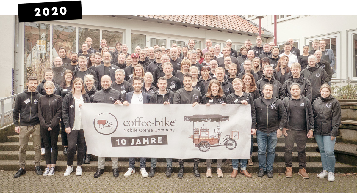 Un grup de angajați Coffee-Bike și parteneri de franciză se află pe o scară cu un banner aniversar de 10 ani și cu data 2020 scrisă cu litere albe deasupra imaginii pe o bară neagră.
