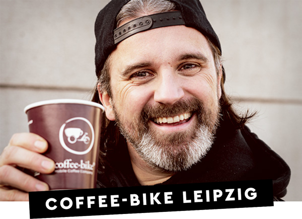 Ein lächelnder Coffee-Bike Franchisepartner hält einen To-go Becher auf dem das Coffee-Bike Logo zu sehen ist und auf einem schwarzen Balken steht in weißer Schrift Coffee-Bike Leipzig 