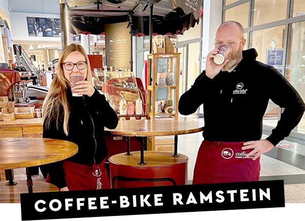 Two Coffee-Bike franchise partners in company clothes with red aprons drink coffee from brown to go cups at their mobile coffee bar in a building and Coffee-Bike Ramstein is written in white on a black bar.
