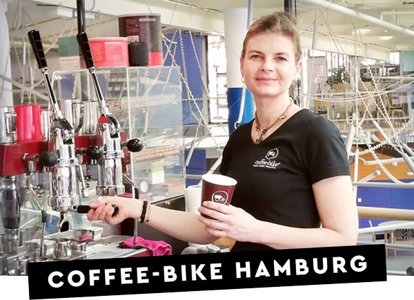 O parteneră zâmbitoare a francizei Coffee-Bike stă cu o ceașcă maro to go în mână la aparatul portafilter al barului său mobil de cafea pentru a trage un espresso, iar pe o bară neagră scrie Coffee-Bike Hamburg cu litere albe.