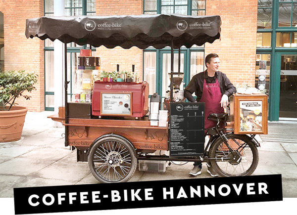 Un partener de franciză Coffee-Bike este gata de acțiune în spatele barului său mobil de cafea din fața unei clădiri, iar pe o bară neagră scrie Coffee-Bike Hanovra cu litere albe.