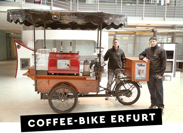 Doi parteneri de franciză Coffee-Bike sunt gata de acțiune lângă barul lor mobil de cafea, iar pe o bară neagră este scris cu litere albe Coffee-Bike Erfurt