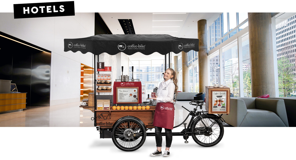 A barista in corporate attire stands smiling with a coffee to go mug in front of a coffee bike in side view against a background showing a hotel reception area with the word hotels in the upper left corner