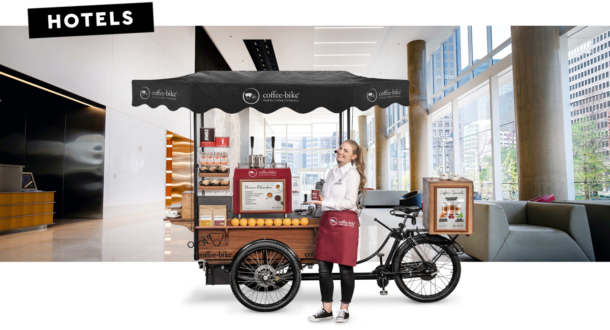 Ein Barista in Firmenkleidung steht lächelnd mit einem Kaffee To-go Becher vor einem Coffee-Bike in Seitenansicht vor einem Hintergrund in dem eine Hotel Empfangsbereich zu sehen ist mit dem Wort Hotels in der oberen linken Ecke