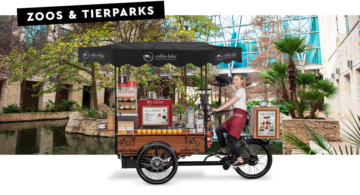 Ein Barista in Firmenkleidung sitzt lächelnd auf einem Coffee-Bike in Seitenansicht vor einem Hintergrund in dem ein Zoo Gehege zu sehen ist mit den Worten Zoos und Tierparks in der oberen linken Ecke