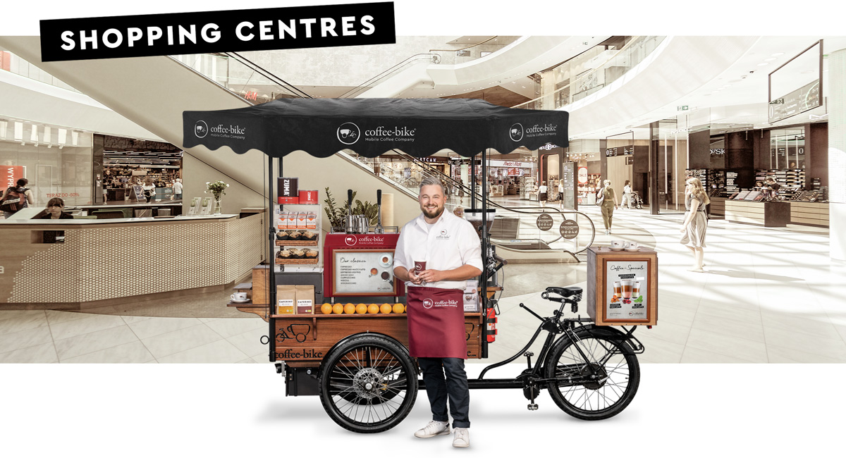 A barista in corporate attire stands smiling with a coffee to-go mug in front of a Coffee-Bike in side view against a background in which the interior of a shopping mall can be seen with the word shopping centres in the upper left corner