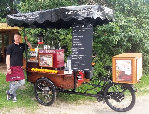 A man in a black T-shirt and red apron leans on the left side of a Coffee-Bike, which stands in front of a green bush.