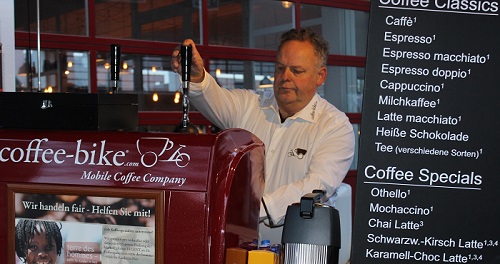 A man in a white Coffee-Bike shirt operates the portafilter machine, which is in focus next to the black beverage board