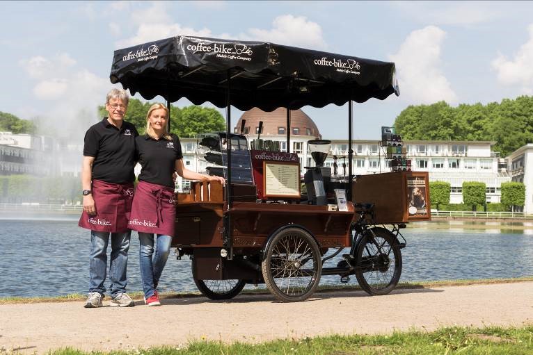 A woman and a man in Coffee-Bike clothes are standing on the left side of a Coffee-Bike, which is standing on a path in front of a river
