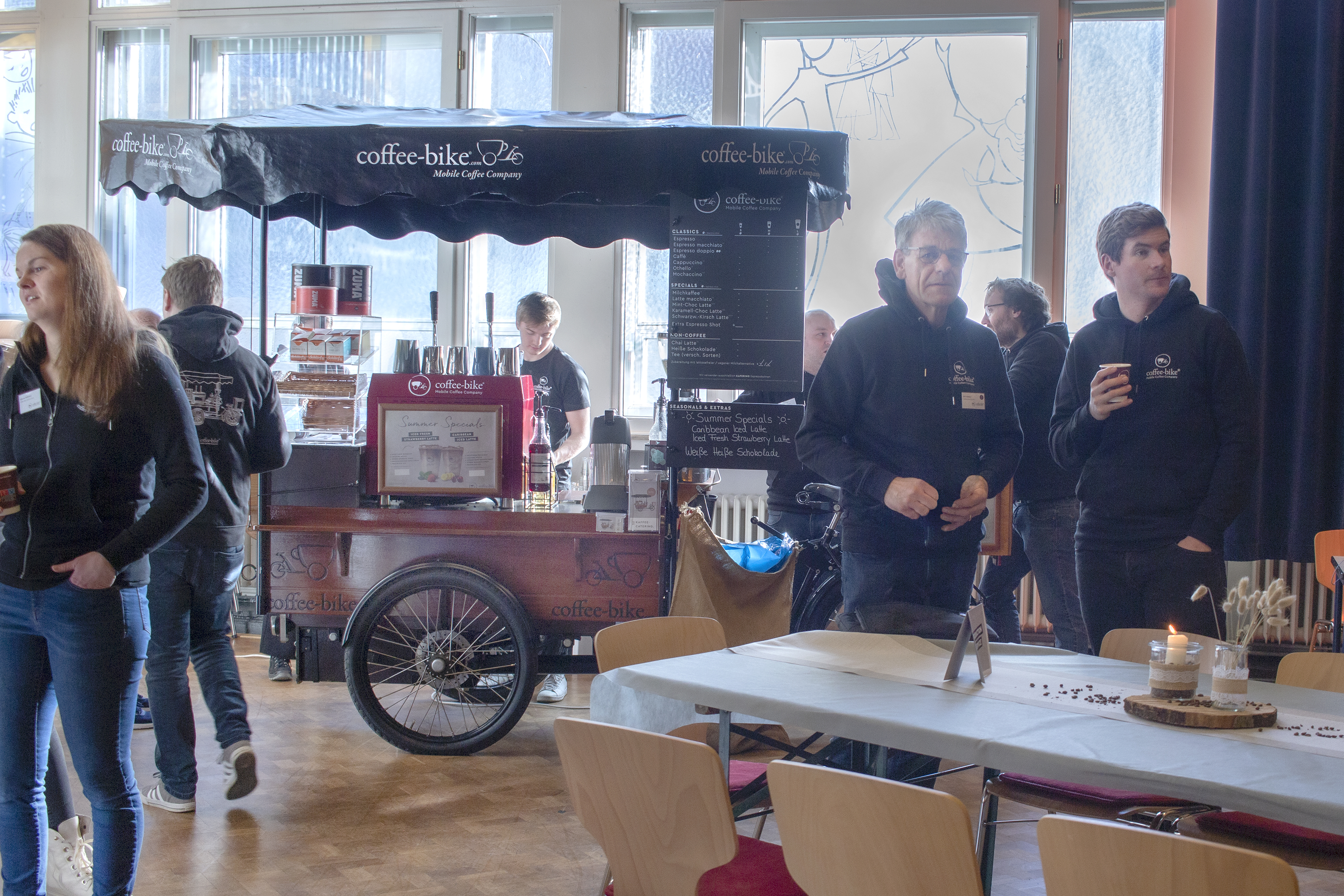 A fully equipped Coffee-Bike stands in a room with tables, chairs and people